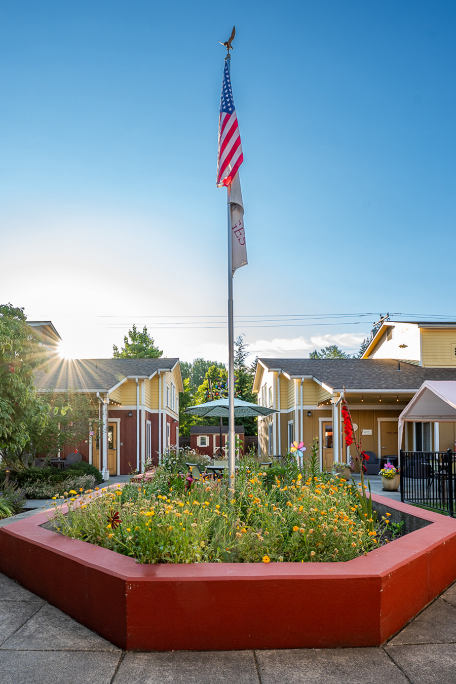 Flagpole surrounded by vibrant flowers and buildings, sunny day.